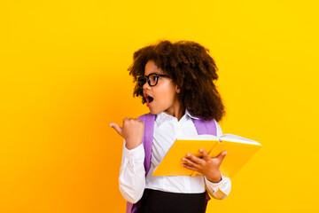 Excited Schoolgirl Holding a Notebook While Pointing with Enthusiasm Against a Vibrant Yellow...