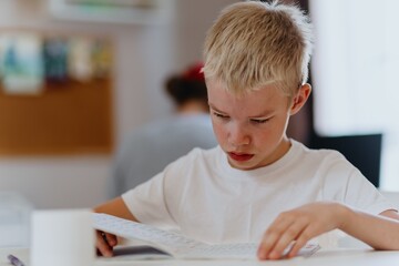 A boy with hearing aids concentrates on homework at a sunlit desk, embodying joyful learning and...