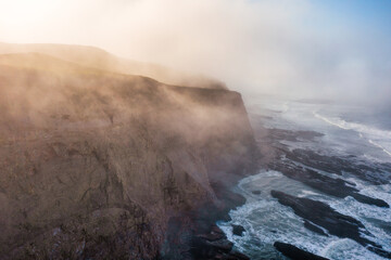 Fog drifts over dramatic cliffs and the rocky shoreline of Scott Creek Beach in California. Aerial...