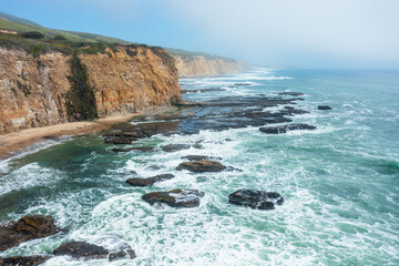 Aerial view of Californias rugged coastline along Highway 1, with steep cliffs, turquoise waters, and sea caves under a bright sky, capturing the wild beauty of the Pacific shore