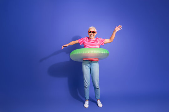 Elderly woman enjoying leisure wearing stylish casual outfit with inflatable ring on vibrant purple background, depicting summer fun - Powered by Adobe