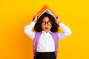 Excited schoolgirl wearing glasses holding an open book as a hat against a yellow background