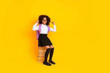 Cheerful schoolgirl with curly hair posing in school uniform, holding a backpack on a yellow background
