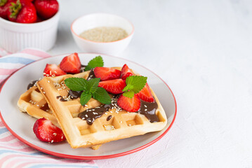 Belgian waffles with strawberries and chocolate topping on the table,  with ingredients in the background
