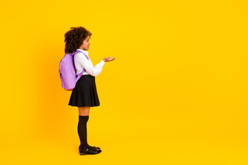Side profile of young schoolgirl in uniform with backpack open hand gesture bright yellow background education concept