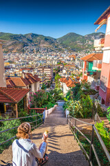 Woman sitting on stairs eating street food, overlooking colorful Mediterranean town and mountains at sunset