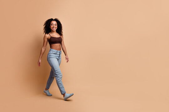 Graceful young woman walking confidently in casual attire with a smile and stylish hair against a beige background
