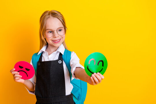 Cheerful schoolgirl holding happy and sad smiley faces for emotion study on vibrant yellow background