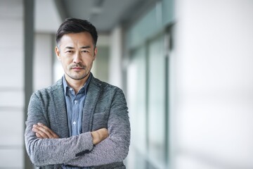 Close-up of a Chinese businessman in smart casual attire, arms crossed in a modern office hallway, captured with diffused studio lighting perfect for technology leadership and business success.