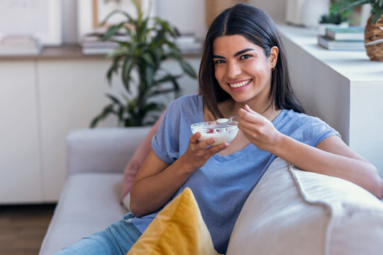 Beautiful woman eating yogurt with strawberries while sitting on the couch at home - Powered by Adobe