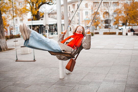 Smiling mature senior woman 55-59 year old rocking on swing at city street wearing vision glasses outdoor. Senior adult lifestyle.