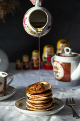 Pancakes with walnuts and honey on rustic breakfast table. Syrup being poured from ceramic jug.