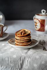 Stack of golden pancakes with walnuts and syrup on ceramic plate. Vintage tea set in background. Cozy morning breakfast scene.