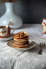 Stack of golden pancakes with walnuts and syrup on ceramic plate. Vintage tea set in background. Cozy morning breakfast scene.