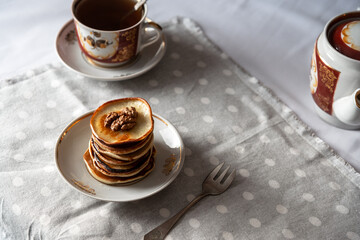 Stack of golden pancakes with walnuts and syrup on ceramic plate. Vintage tea set in background. Cozy morning breakfast scene.