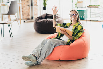 Businesswoman woman relaxing on bean bag chair while studying, holding notebook and waving in a...