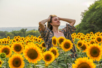 young blond woman among sunflowers in a field, doing her hair, straightening her hair with her...