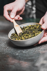Close-up of hands serving dried tea leaves from a bowl with a wooden scoop, highlighting texture...