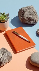 A minimalist flat lay featuring a succulent plant, a textured orange notebook, a pen, and various natural stones on pastel blue and peach backgrounds.