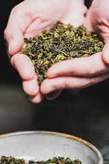 Hands holding loose dried green tea leaves above a ceramic bowl. Natural texture and organic look,...