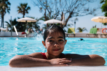 A young girl is in a pool, smiling and looking at the camera. The scene is bright and cheerful, with several umbrellas and chairs around the pool area