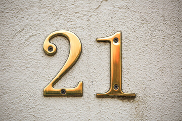 Shiny brass house numbers 21 affixed to a light-colored, textured wall. The digits are clean and...