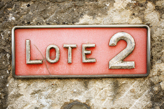 A weathered metal sign reading "LOTE 2" mounted on a rough stone wall. The red background and aged, raised silver letters give the sign a rustic, vintage look in a natural outdoor setting