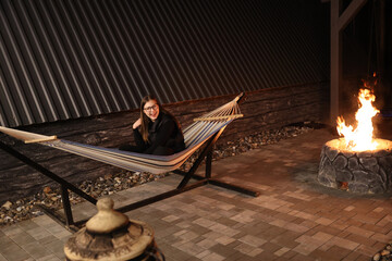 A young woman relaxes on a striped hammock by a cozy firepit at night in a pine forest. Peaceful...