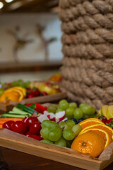 Colorful wooden tray filled with fresh fruits and vegetables: green grapes, oranges, cherry tomatoes, cucumbers, radishes, and berries. Ideal for healthy eating, catering, vegan lifestyle