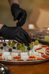 Chef hands in black gloves preparing elegant appetizers: skewers with grapes and blueberries, soft brie cheese, and tartlets with red caviar. Perfect for catering, gourmet cuisine, or culinary service