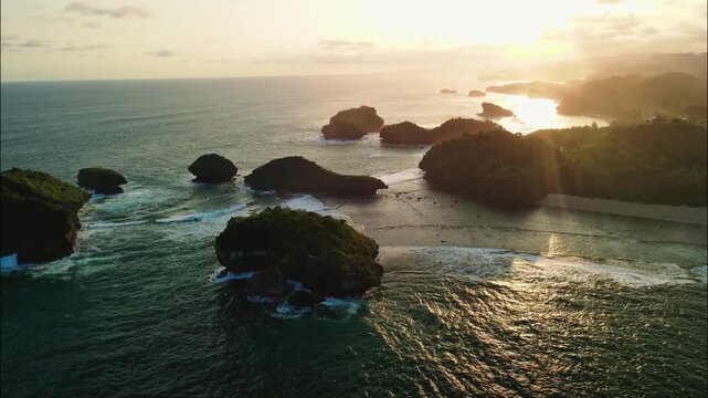 A stunning seascape unfolds with numerous green-topped sea stacks and rocky islands scattered across the ocean, all bathed in the warm, hazy light of sunset. Watu Karung Beach, Pacitan.