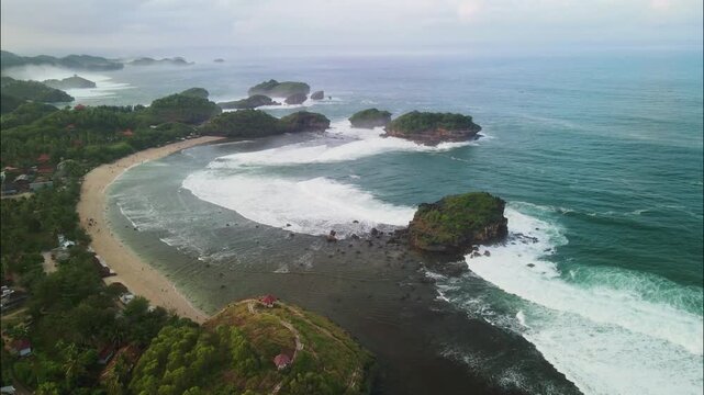 From a cliff-top viewpoint, this stunning aerial shot captures a crescent beach and a bay dotted with numerous green, rocky sea stacks and crashing waves. Aerial Watu Karung beach in Pacitan.