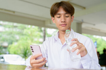 Non binary university student sitting in college campus drinking water and using phone