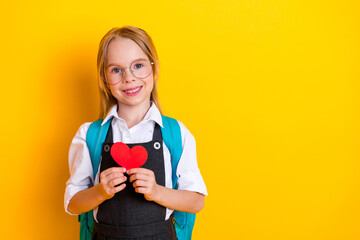 Happy schoolgirl holding a red paper heart, standing against a bright yellow background, wearing a...