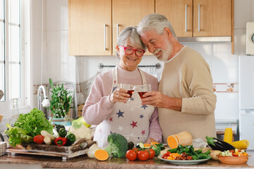 Cheerful senior couple toasting with red wine glasses working together in the home kitchen...