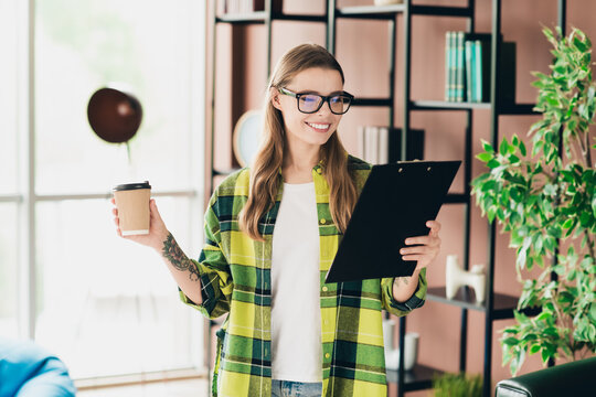 Successful professional woman reviewing documents indoors, holding coffee cup, enjoying coworking space office ambiance