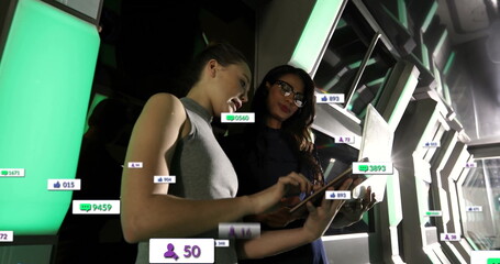 Two women with glasses tracking metrics in futuristic corridor, with tablet, laptop, floating icons