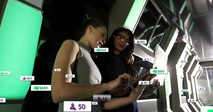 Two women with glasses tracking metrics in futuristic corridor, with tablet, laptop, floating icons