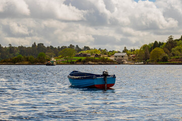 Fototapeta premium Empty blue boat sits on river with rocky shore with houses in background in West Cork region, Glengarriff, Ireland