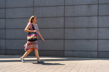 Mid adult woman walking against background of gray modern wall in summer. Copy space. Side view