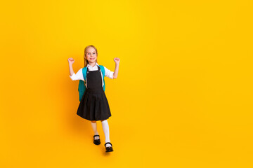 Smiling schoolgirl in uniform with backpack standing on yellow background, expressing joy for back-to-school season