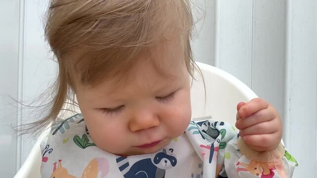 Adorable baby girl in colorful bib sitting in a high chair, exploring food with curiosity while practicing baby led weaning with fruits, vegetables, and eggs during an outdoor mealtime at home