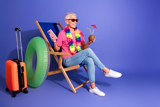 Stylish senior woman in summer outfit enjoying leisure moments with tropical drink and travel accessories on a vibrant purple background