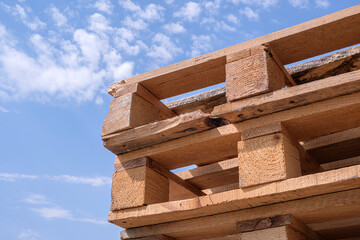 Close-up view of several rough wooden pallets stacked outdoors under a bright blue sky with scattered clouds. Highlighting rustic industrial design and recycling themes.