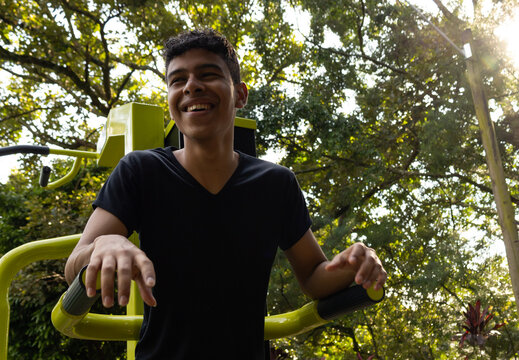 Cheerful young Latino man doing strength training, doing dips on parallel bars in a park in the morning on a sunny day, healthy lifestyle outdoors