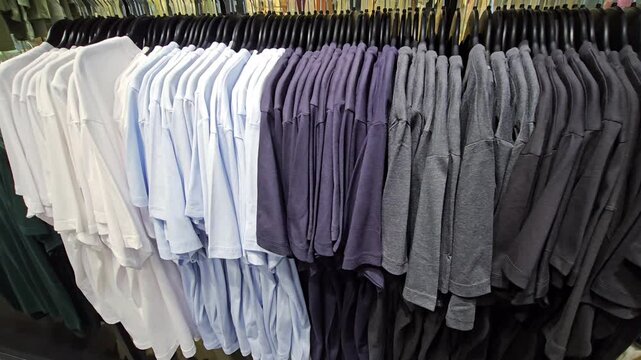 Row of plain t shirts hanging on rack in clothing store arranged by color from white to black, retail display of casual apparel on hangers inside shop interior wardrobe selection and shopping concept