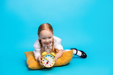 Smiling young schoolgirl lying on a cushion with an alarm clock in a bright background symbolizing...
