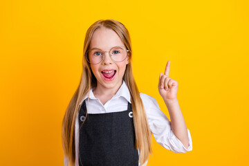 Excited young schoolgirl gesturing with her finger up in front of yellow background promoting...