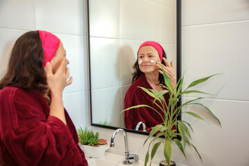50 year old mature woman looking at her wrinkles in the mirror and taking care of her skin with...