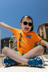 Smiling child wearing sunglasses and an orange T-shirt sitting outdoors on a sunny day in Calpe,...